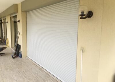 Volet roulant blanc complètement fermé sur une baie vitrée en façade d’un appartement avec terrasse carrelée.